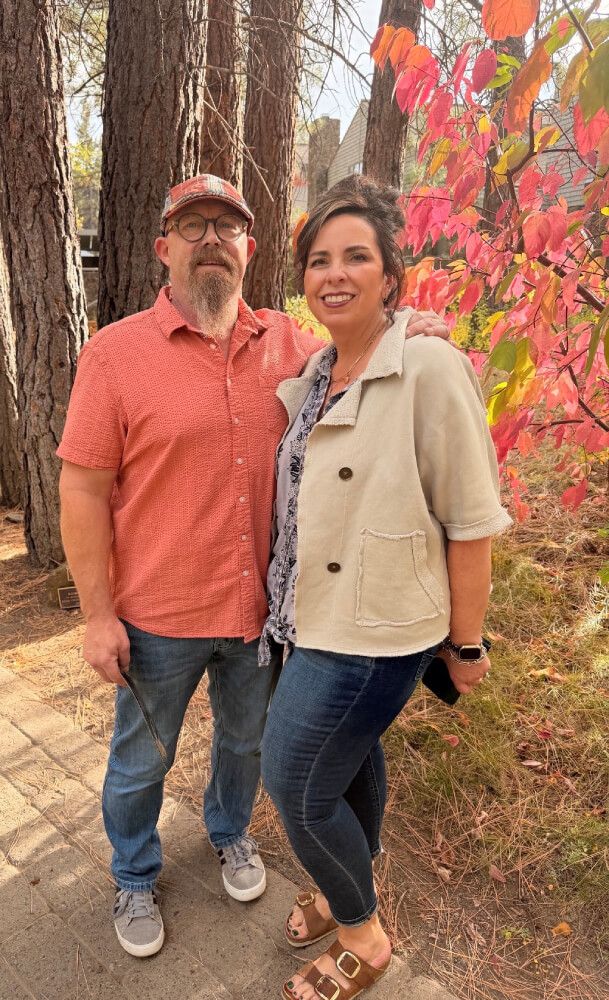 A couple standing outdoors among red and orange autumn leaves, smiling at the camera.