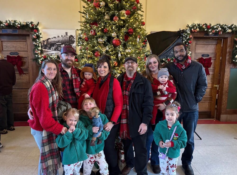 Family group posing in front of a decorated Christmas tree in a festive indoor setting, wearing red and green outfits.